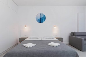A simple bedroom featuring a bed, bedside lamp, and a small table. The design is clean and minimalistic.