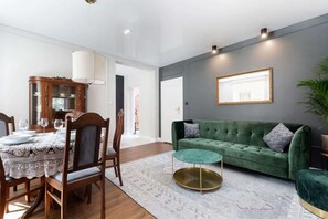 A bright living room with comfortable seating and classic wooden dining furniture. Large windows fill the room with sunlight.