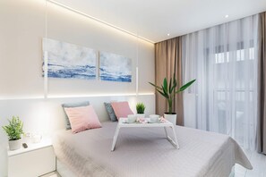 A bedroom with a light color scheme, large windows, and soft, inviting bedding.