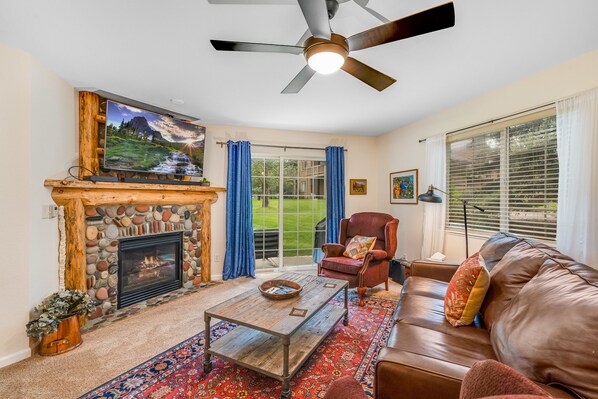 A cozy living room with a ceiling fan, stone fireplace, large window, leather sofa, armchairs, wooden coffee table, and TV mounted above the fireplace. Blue curtains frame a view into a green yard.