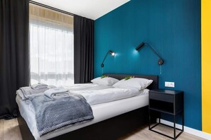 A cozy bedroom with a double bed featuring white linens. A window offers a view of the outside, with heavy curtains for privacy.
