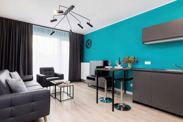 A modern living room featuring a turquoise accent wall, a gray sofa bed, and a sleek dining table with chairs. The room is brightly lit by natural light from a large window.