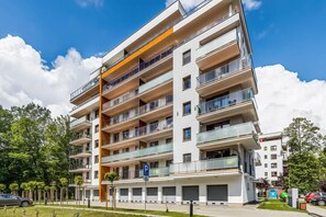 A modern multi-story building with balconies, surrounded by greenery and a clear blue sky.
