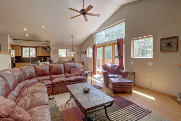 A spacious living room with high ceilings, a large brown leather sectional sofa, a matching armchair, a coffee table, and an open kitchen with bar stools in the background. Large windows let in natural light.