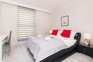 A bedroom with a double bed, dressed in white and gray bedding, with red  accent pillows. The room includes nightstands with small lamps, and a large window with modern blinds for natural light.