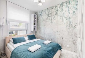 A bedroom with a double bed, teal bedding, and an accent wall with a floral or artistic design. The room appears well-lit and inviting.