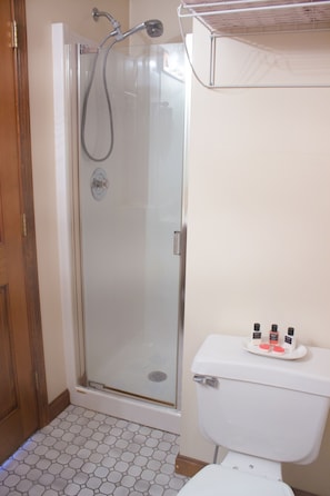 Standup shower with hand-held nozzle and toilet topped with starter toiletries. 