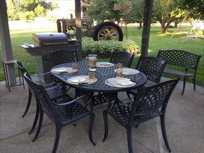 A beautifully set backyard dining area featuring a spacious table with six comfortable chairs and a charming bench, perfect for gathering with family and friends. Nearby, a sleek BBQ grill stands ready for sizzling steaks and fresh-off-the-grill delights.