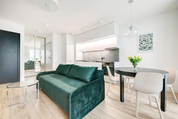 A modern living room featuring a green velvet sofa, with a minimalist design. There's a dining area in the background.