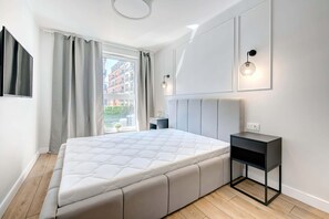 A cozy bedroom with a large bed, white walls, and soft natural light from the window.