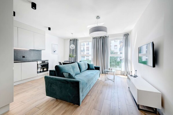 A bright, modern living room with a blue sofa, large windows, and an open plan layout leading to the kitchenette.
