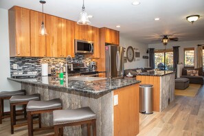 Modern kitchen and living area with wooden cabinets, granite countertops, stainless steel appliances, an island with barstools, a ceiling fan, and a wall-mounted dartboard.