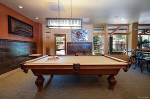 A room with a pool table under a rectangular light fixture, artwork on the walls, tile and carpet flooring, and large windows providing natural light.