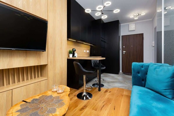 A cozy living room with a modern kitchenette. It features sleek black cabinets, a wooden countertop, and decorative shelves.