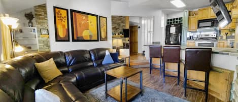 A living room with a curved black leather sectional sofa, a wooden coffee table, three bar stools at a kitchen counter, wall art, and a ceiling fan. The kitchen features stainless steel appliances.