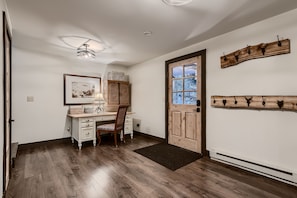 A small entryway features a wooden desk with a chair, a framed artwork above the desk, and a door with a window. Two coat racks are mounted on the wall next to the door.