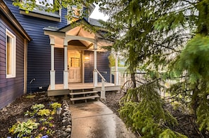 A blue house with a white porch, lit by a hanging lantern, and a concrete pathway lined with plants and rocks, leading to the front door.