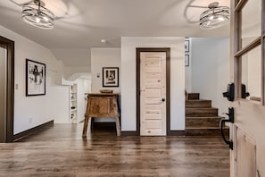Modern interior hallway featuring wooden flooring, a wooden door, staircase, framed artwork, and a wooden console table under contemporary ceiling lights.