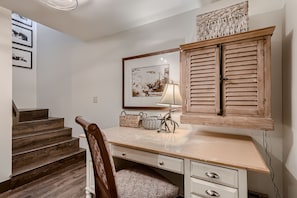 A workspace with a wooden desk, a cushioned chair, a lamp, woven baskets, and wall art. Steps to the left lead upwards.