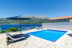 A spacious swimming pool with comfortable deckchairs and a parasol and a view of the crystal blue sea from the property of a luxury villa for a family holiday on Korčula