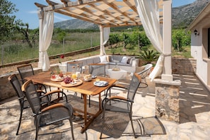 The stone-paved courtyard of villa Zlatni Rat in Bol with a canopy, a dining table and a set for relaxing, surrounded by a vineyard and olive trees.