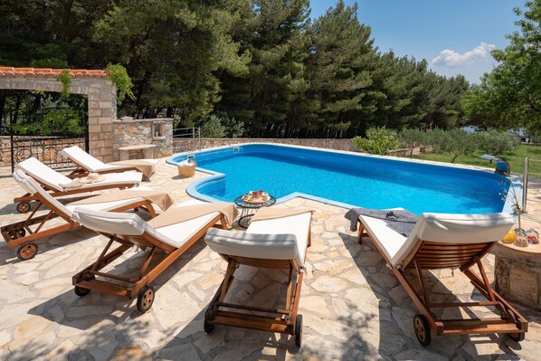 The stone terrace of the Dalmatian villa in Bol with sea view, 6 deckchairs with mattresses and towels, a pool with a waterfall. The property has pine and olive trees.