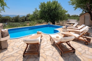 Sunbathing stone terrace of the Dalmatian villa Zlatni Rat in Bol with a view over the sea, olives and almond trees. 6 sunbeds with mattresses by the pool with a waterfall.
