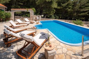 The stone terrace of the Dalmatian villa in Bol with sea view, 6 deckchairs with mattresses and towels, a pool with a waterfall. The property has pine and olive trees.