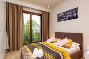 A bedroom featuring a queen-size bed with vibrant yellow and gray bedding. A large glass door leads to the balcony, allowing natural light to fill the space.

