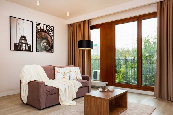 A cozy living room with a sofa, a coffee table, and a TV mounted on the wall. Large windows with wooden frames provide plenty of natural light and offer a view of the greenery outside.