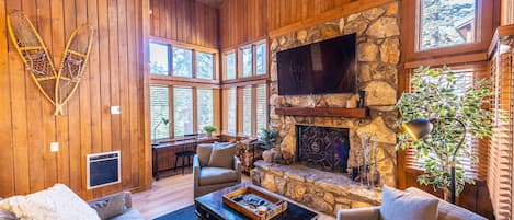 Living room w/ 30ft ceilings, wood fireplace, 65” Smart TV, cozy sofa, desk area