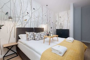 A cozy bedroom with a double bed adorned with white and yellow bedding, a decorative floral wallpaper, and minimalistic bedside tables with lamps.