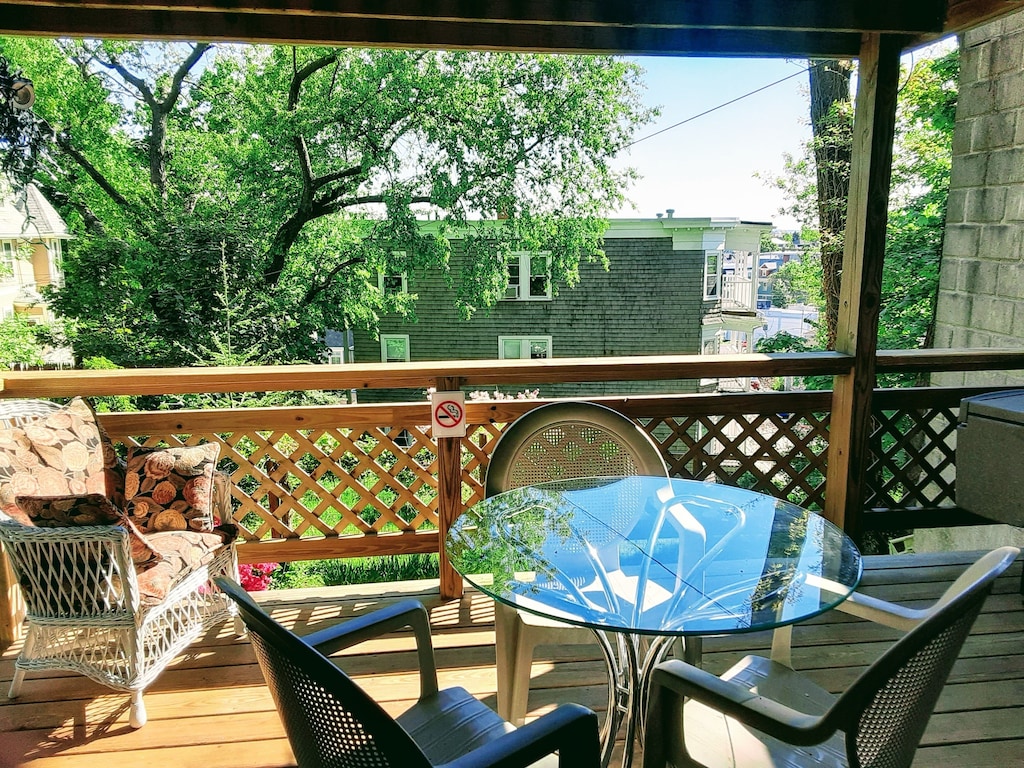 A vacation rental in Boston has a sun soaked terrace with whicker furniture, a table and chairs, and views of a large tree.
