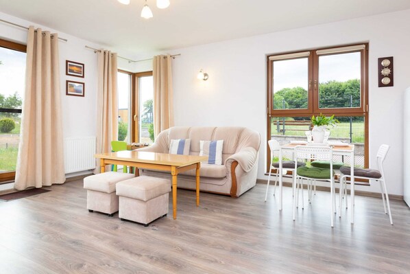 A bright living room with large windows, a comfortable sofa, a coffee table, and a dining area with a wooden table and chairs.
