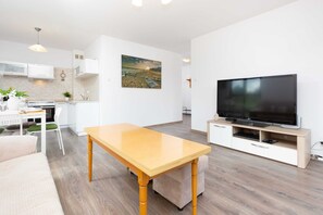 A bright living room with large windows, a comfortable sofa, a coffee table, and a dining area with a wooden table and chairs.