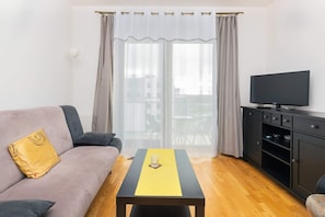 A living room with a comfortable gray sofa, yellow cushions, and a flat-screen TV. The natural light adds warmth to the space.