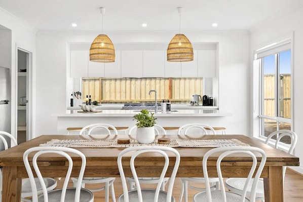 The stunning fresh white kitchen has wicker pendant lights over the entertainer's island bench which takes centre stage in this beautiful home. There are bar stools for informal meals and a dining table for 8 guests. 