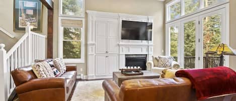 The stunning living room boasts high ceilings, picture windows, gorgeous built-ins surrounding the gas fireplace, a flat-screen TV,  coffee table, cozy armchair, end table, lamp and access to the outdoor deck with a gas BBQ grill.