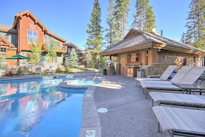 Outdoor pool with a hot tub surrounded by lounge chairs and a small shaded cabana area, set next to mountain-style buildings and tall trees.