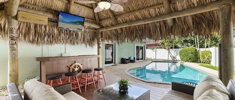 Tropical tiki hut with outdoor bar, tv, and lounge area overlooking heated pool