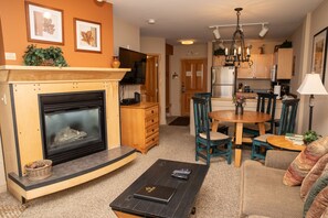 A cozy living area with a fireplace, wooden furniture, a round dining table with four chairs, and a small kitchen in the background. Wall decor includes framed artwork and a chandelier hangs from the ceiling.