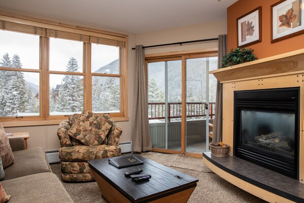 A cozy living room features a beige sofa, floral-patterned armchair, wooden coffee table, and a fireplace. Large windows and a glass door provide a view of a snowy mountain landscape outside.