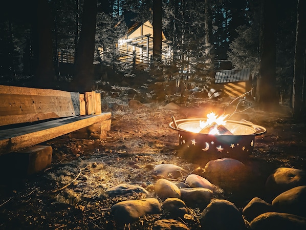 Creekside fire pit and bench under tall pines and starry skies at night