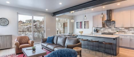 A modern open-concept living room and kitchen featuring a brown leather sofa, red area rug, a central coffee table, bar stools at the kitchen island, and large windows with a snowy outdoor view.