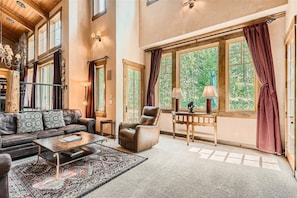 A well-lit living room with a high ceiling, large windows, a dark leather couch, two armchairs, and a wooden coffee table. The space has light walls, a patterned rug, and draped curtains.