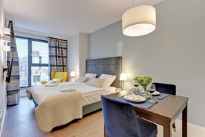 A spacious bedroom featuring a comfortable double bed and a small dining table for two. Large windows provide plenty of natural light.