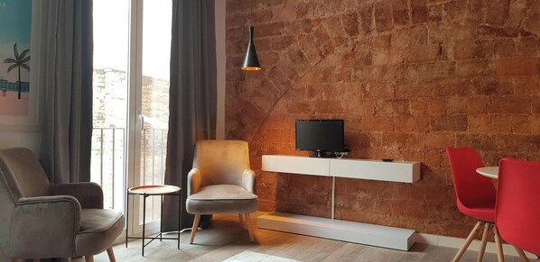 The living room is a modern and cozy space with an exposed brick wall that adds a touch of industrial charm. The room features a large window with grey curtains and a balcony offering natural light. The room is furnished with comfortable seating, includin