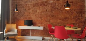 The living room features a modern design with exposed brick walls, a flat-screen television, and a dining table with four bright red chairs. Natural light floods the space, highlighting the exposed brickwork and creating a warm and inviting ambiance.