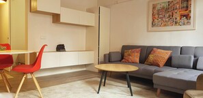 The living room is a bright and airy space with a large, comfortable grey sofa and two pillows. There is a small, round coffee table in the center of the room and a white, tiled floor. Four chairs are positioned at a white table. The room has a minimalist