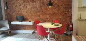 The dining area features a round, white table with red chairs and wooden legs, perfect for enjoying meals together. The exposed brick wall adds a touch of rustic charm to the modern space. The modern, flat-screen television mounted on the white shelf adds